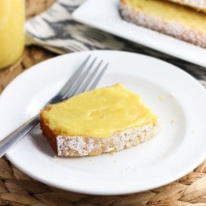 A slice of curd-topped pound cake on a dessert plate with a fork next to the rest of the sliced pound cake and a jar of curd