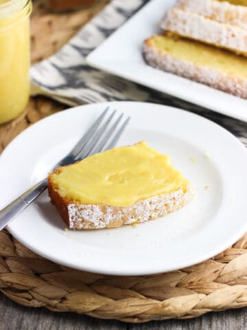 A slice of curd-topped pound cake on a dessert plate with a fork next to the rest of the sliced pound cake and a jar of curd