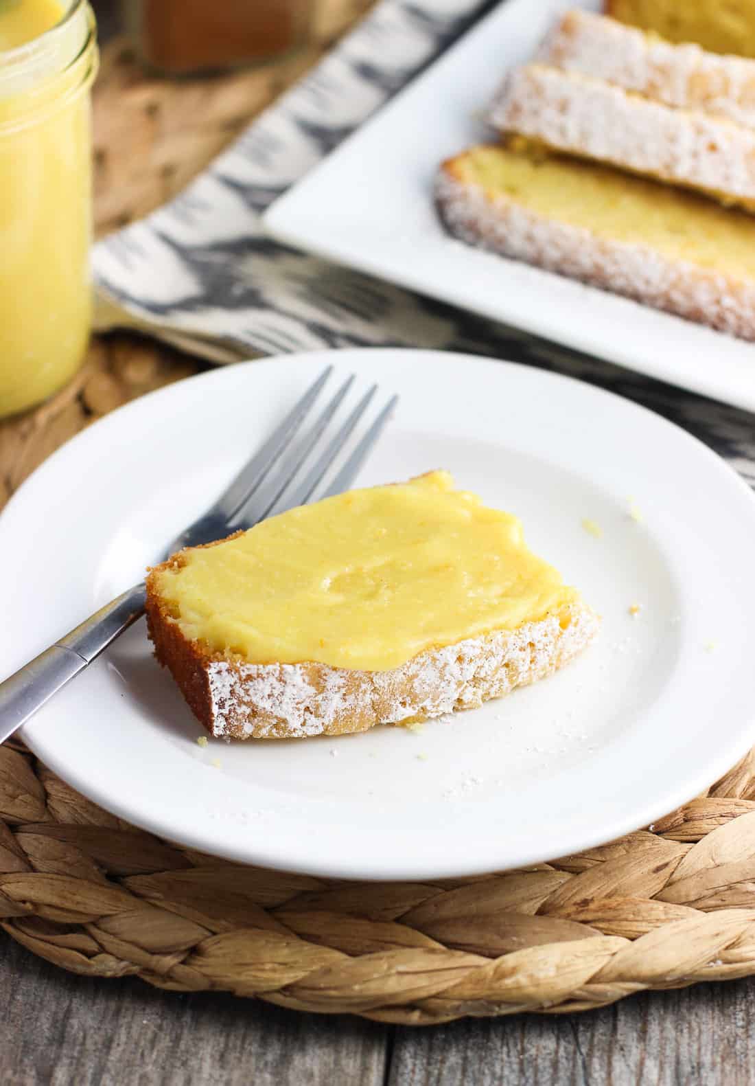 A slice of curd-topped pound cake on a dessert plate with a fork next to the rest of the sliced pound cake and a jar of curd