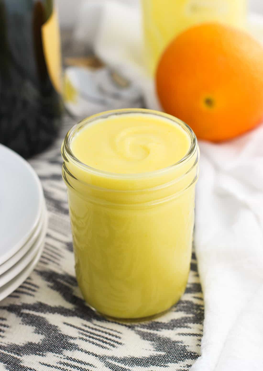 A glass jar filled with orange curd next to a stack of plates, a bottle of champagne, and an orange