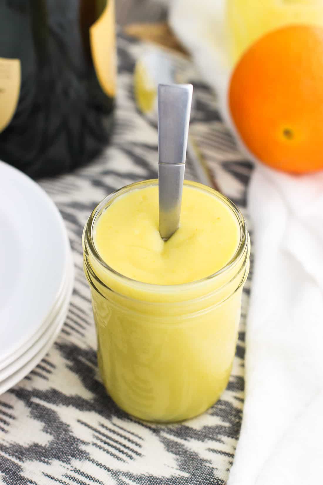 A glass jar of curd with a spoon in the middle next to a stack of plates, a bottle of champagne, and an orange