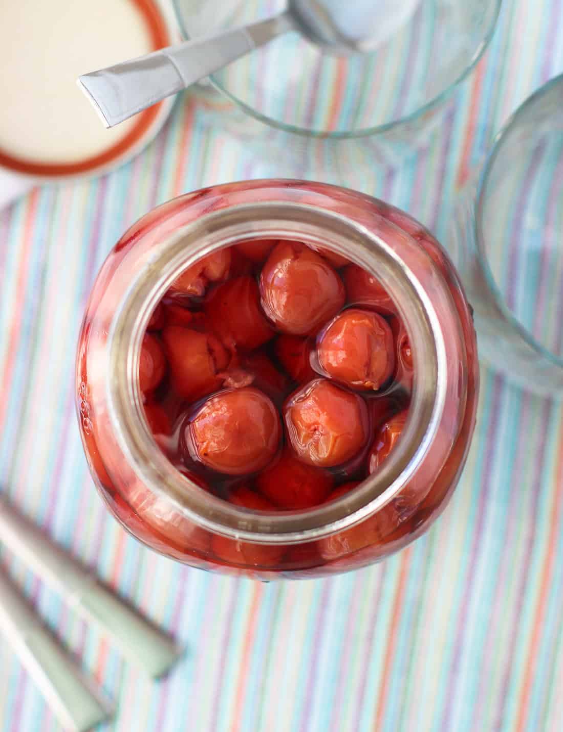 An overhead shot of a large glass jar of homemade maraschino cherries next to an empty rocks glass and spoons