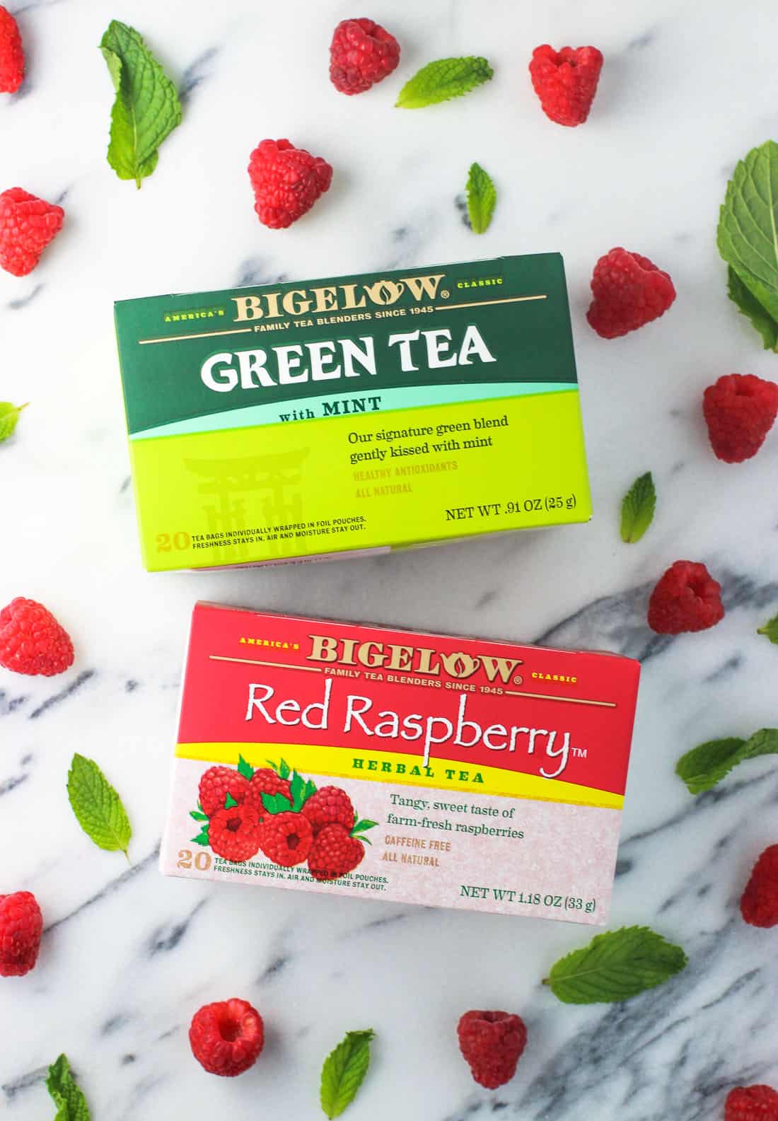 A box of green tea and a box of red raspberry tea on a marble board surrounded by raspberries and mint leaves