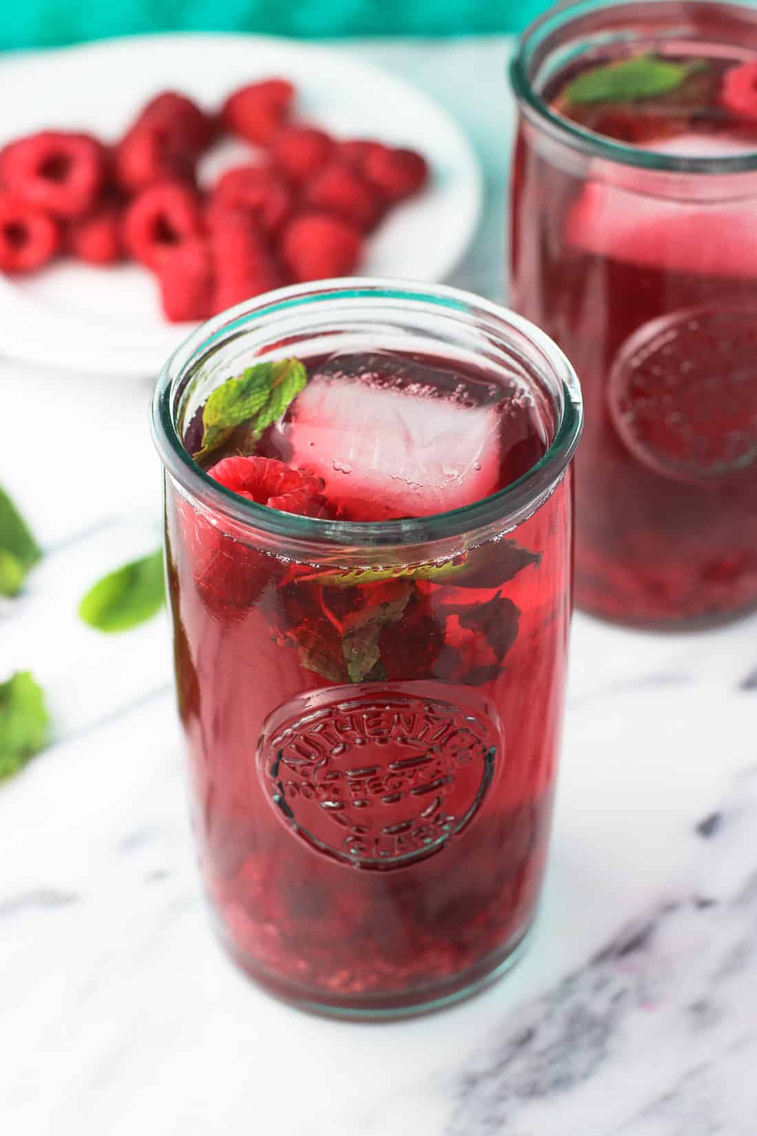 A glass of iced tea with a small dessert plate of fresh raspberries and mint leaves in the background