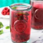 A clear glass of iced mint raspberry green tea on a marble board garnished with raspberries and mint leaves