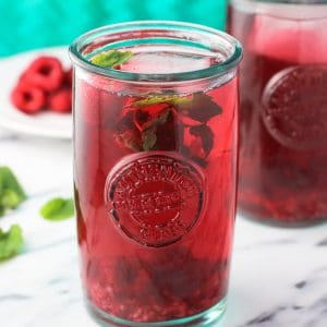 A clear glass of iced mint raspberry green tea on a marble board garnished with raspberries and mint leaves