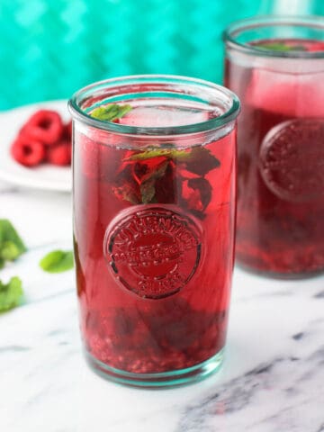 A clear glass of iced mint raspberry green tea on a marble board garnished with raspberries and mint leaves