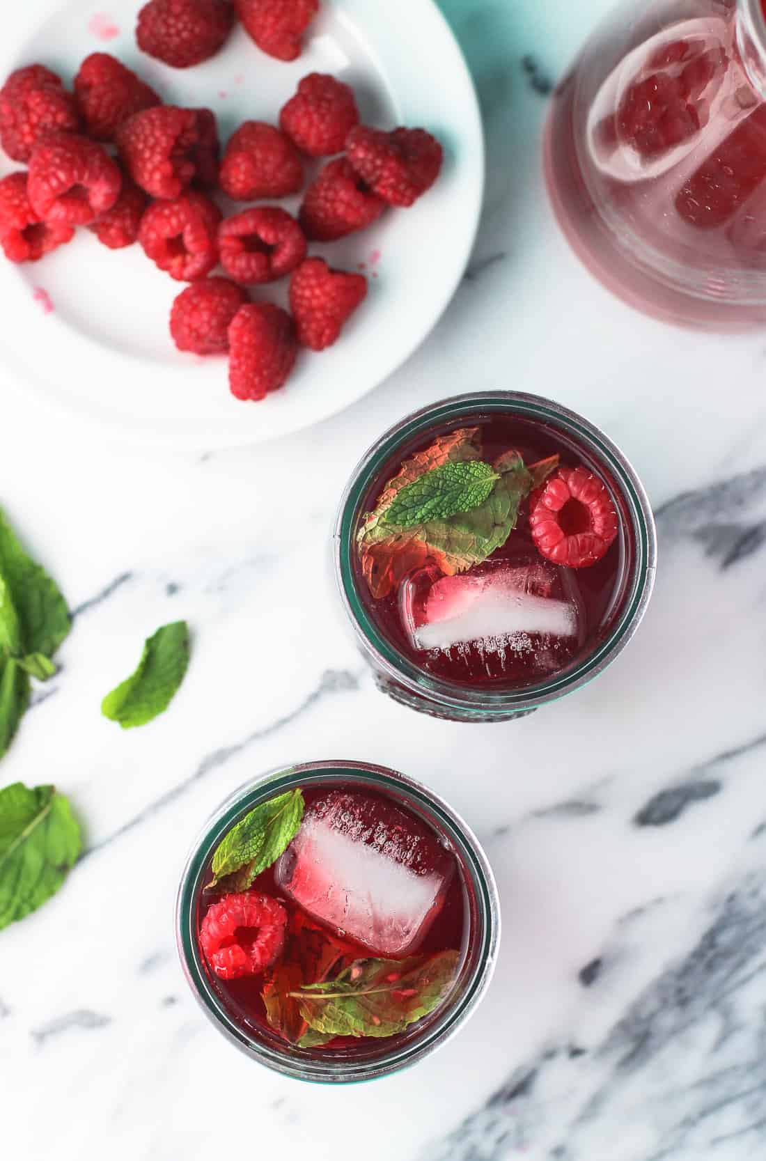 An overhead shot of two glasses of iced tea, raspberries, mint leaves, and a mostly-empty glass carafe of iced tea