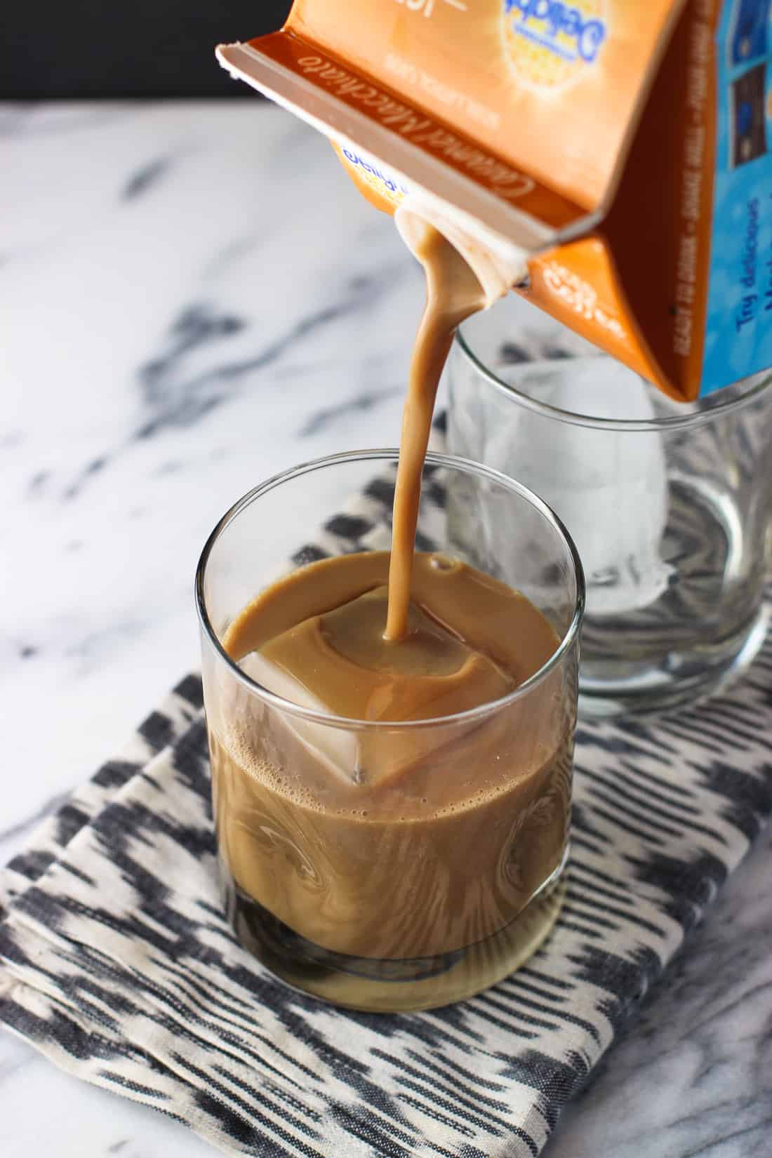 A carton of caramel iced coffee being poured into a small glass with a large ice cube