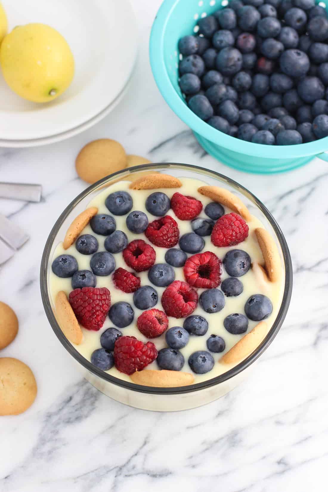 A bowl of lemon pudding made with vanilla wafers and fresh fruit surrounded by recipe ingredients.