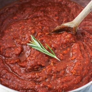 A large pot of tomato sauce with a wooden spoon garnished with a fresh rosemary sprig