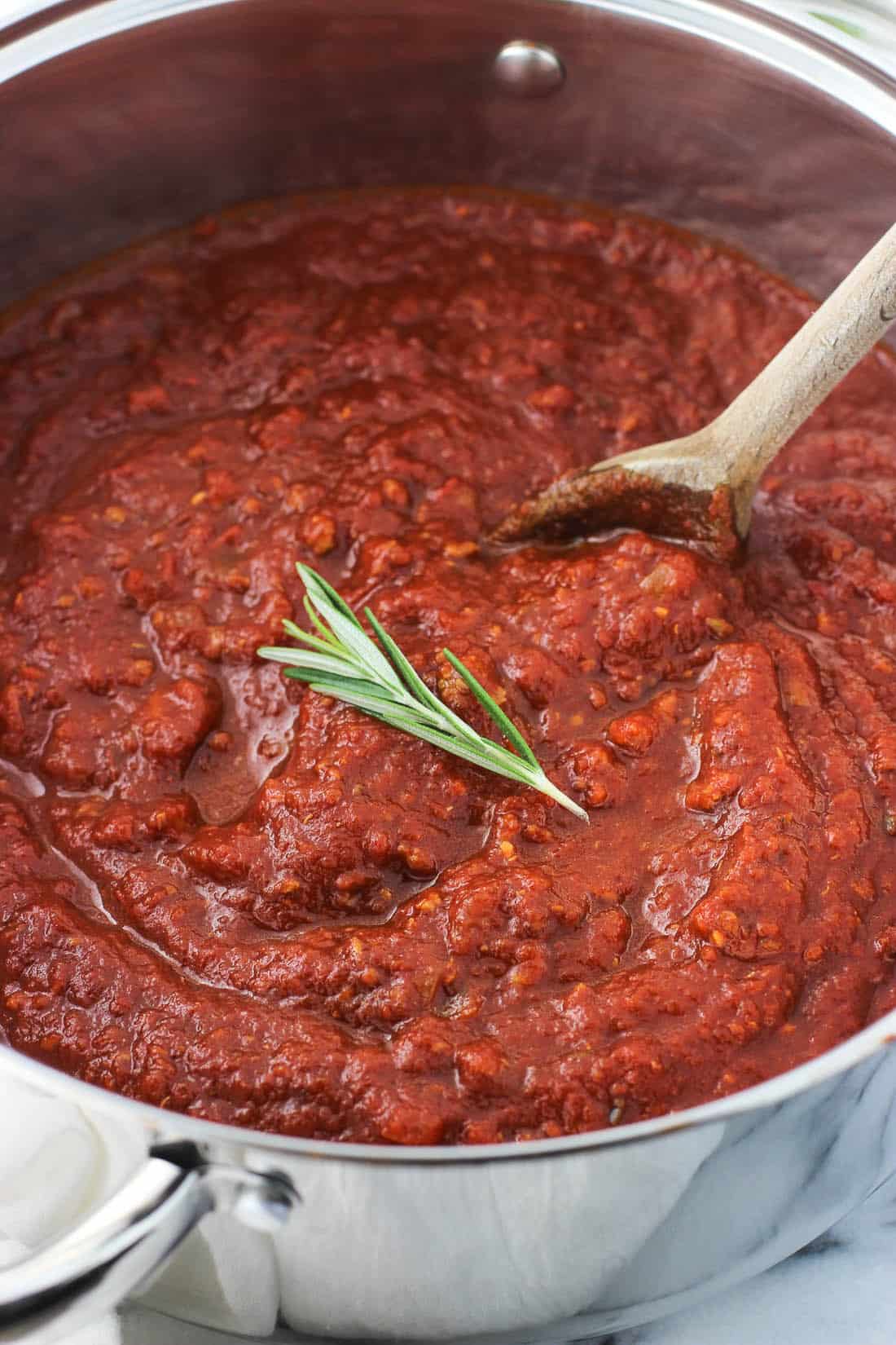 A large pot of tomato sauce with a wooden spoon garnished with a fresh rosemary sprig.