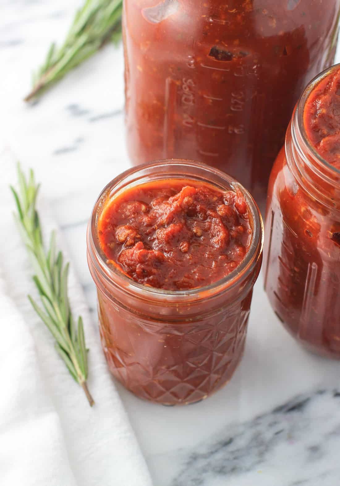 Glass mason jars of various sizes filled with sauce and surrounded by fresh rosemary sprigs.