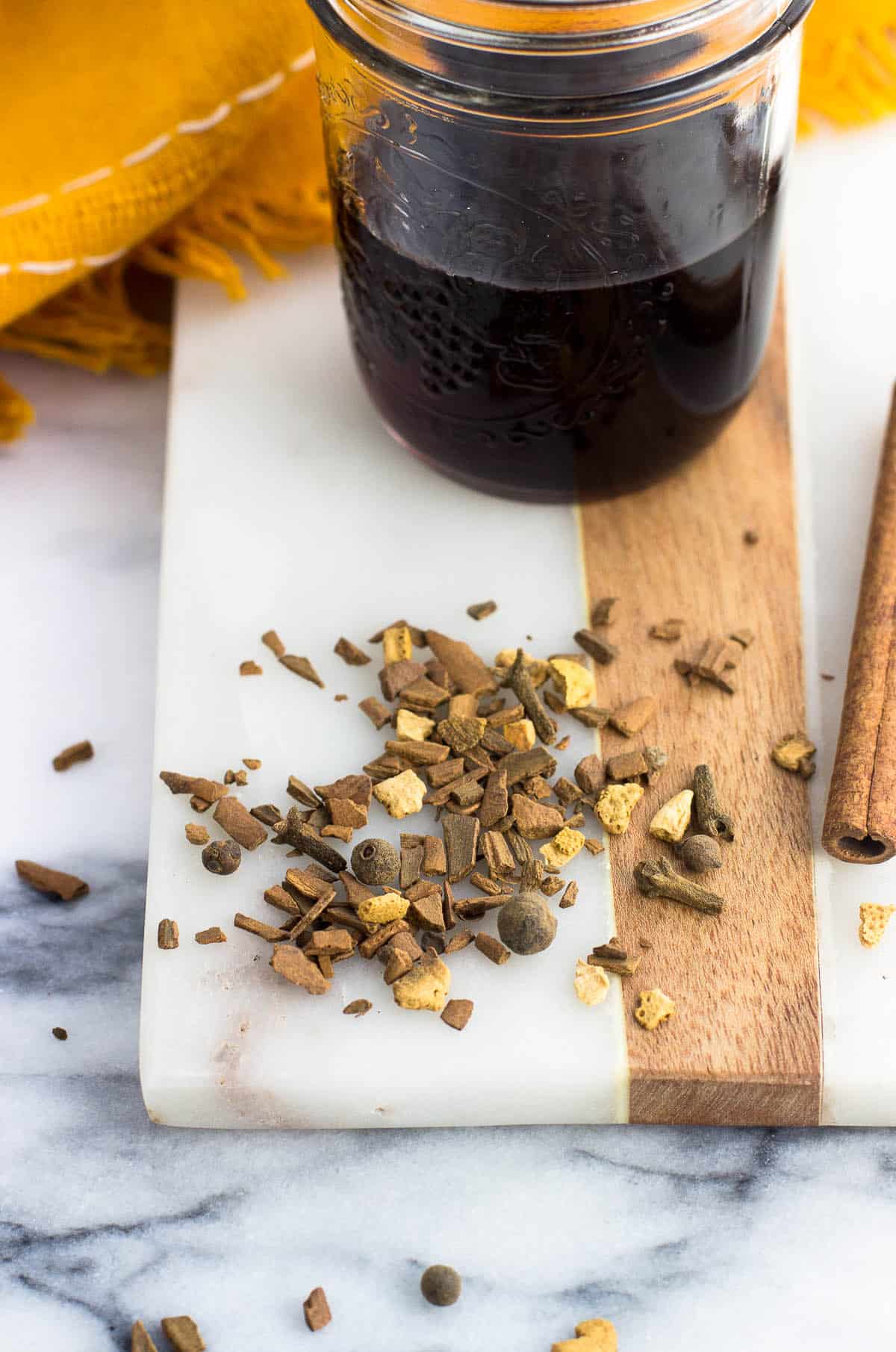 Dried mulling spices scattered on a marble board next to whole cinnamon sticks and a glass jar of wine.