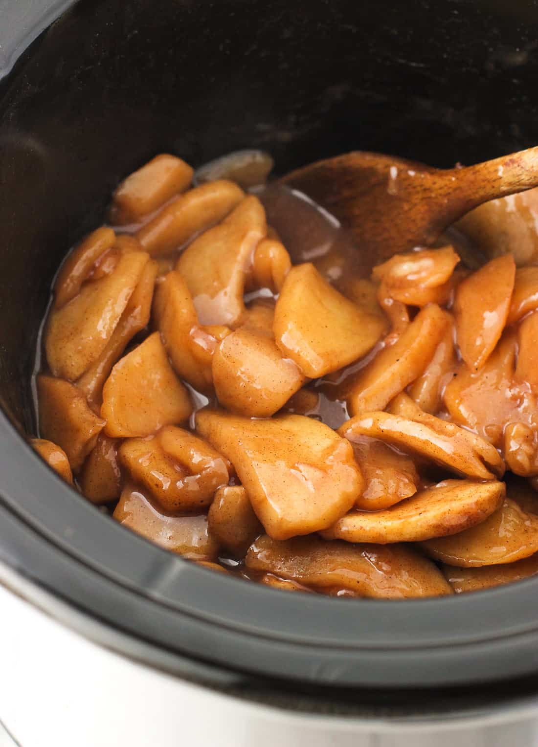 Stewed apple slices in a slow cooker with a wooden spoon.