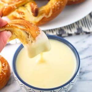 A hand dipping a soft pretzel piece into a bowl of cheese dip.