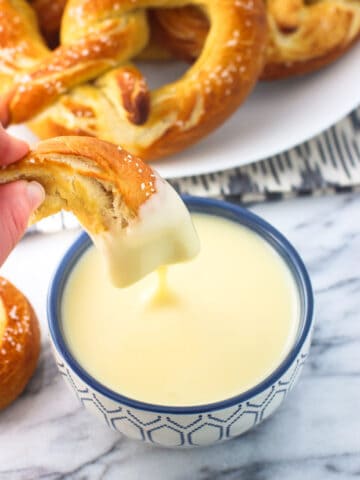 A hand dipping a soft pretzel piece into a bowl of cheese dip.
