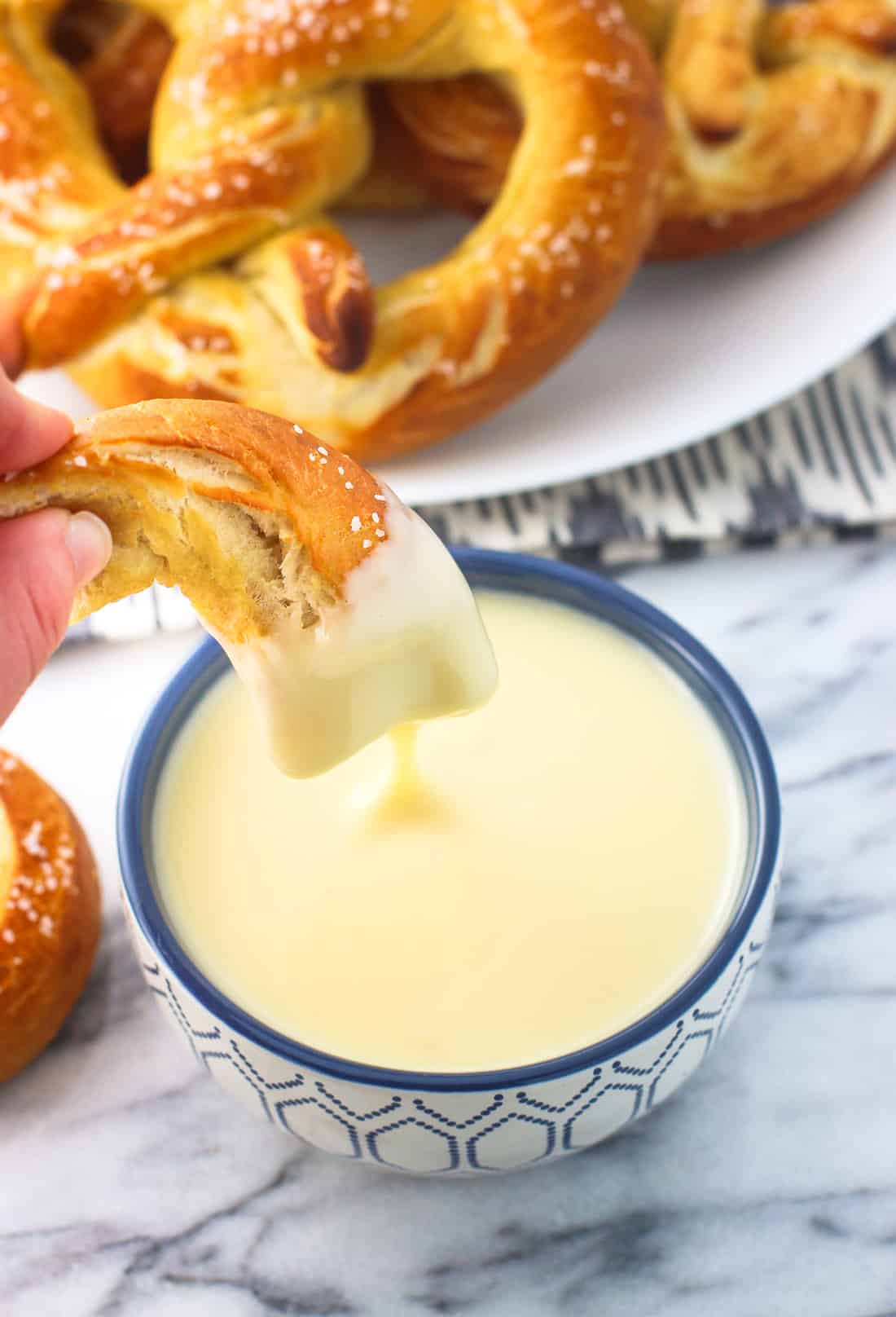 A hand dipping a soft pretzel piece into a bowl of cheese dip.