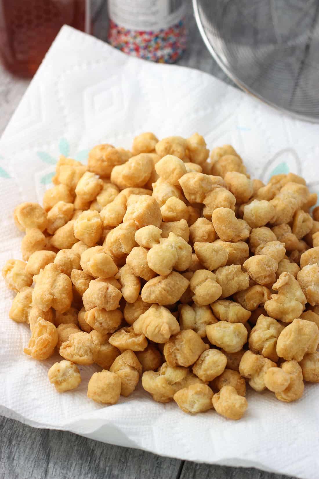 Fried balls of dough on a paper towel in front of a jar of honey, a canister of sprinkles, and a slotted skimmer