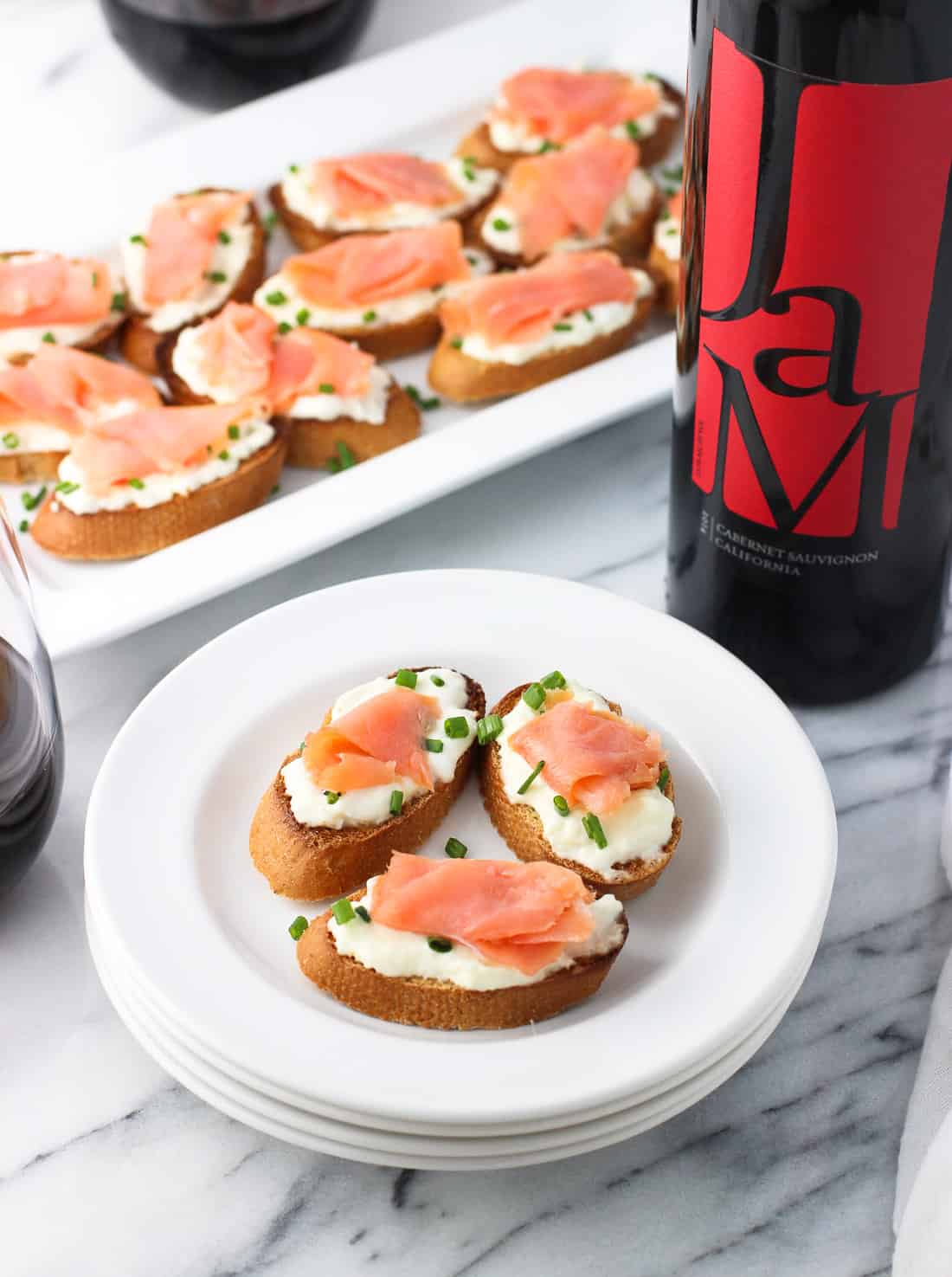 Crostini topped with burrata and a slice of smoked salmon on a plate next to a bottle of wine and the tray.