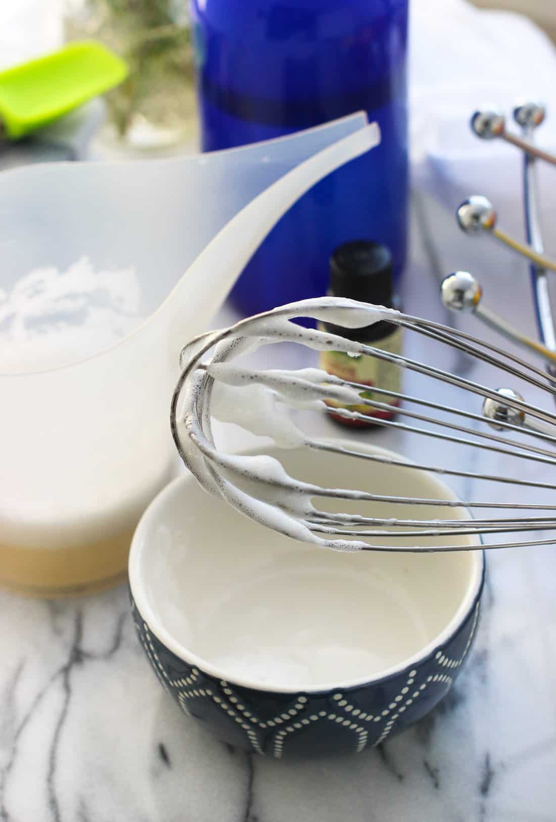 A coated whisk lifted out of a measuring cup of a soap mixture.