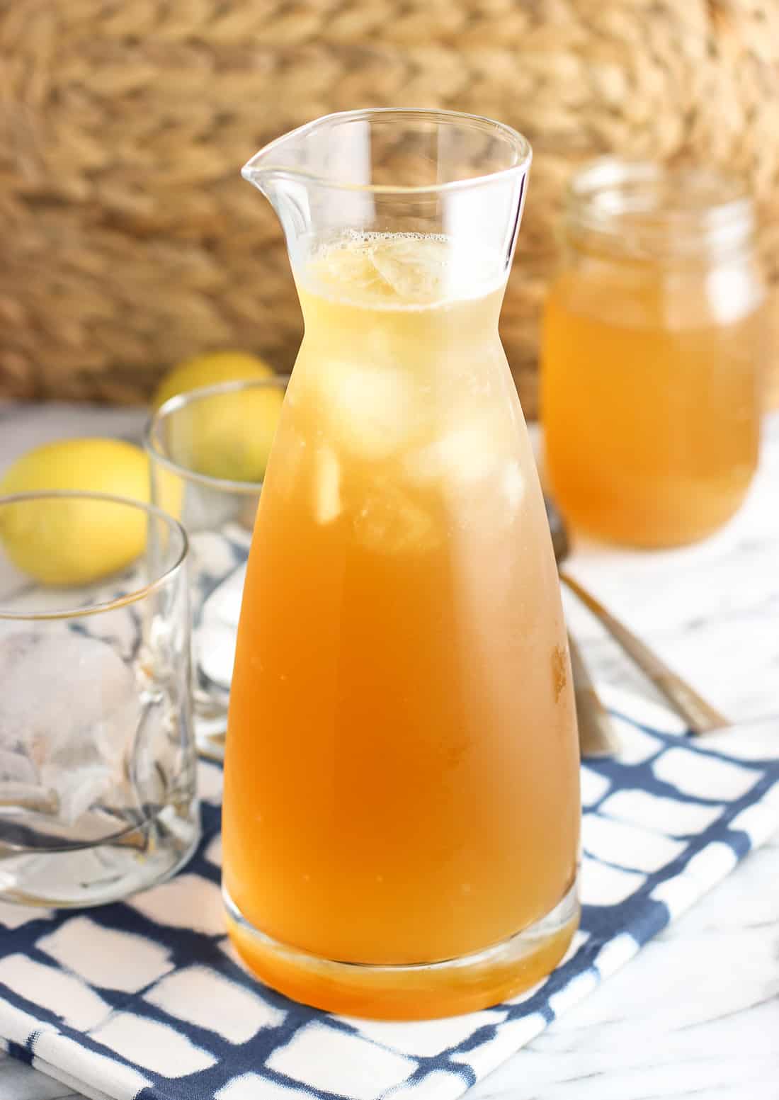 A pitcher of lemon green tea with ice in front of empty glasses, a jar of simple syrup, and two whole lemons