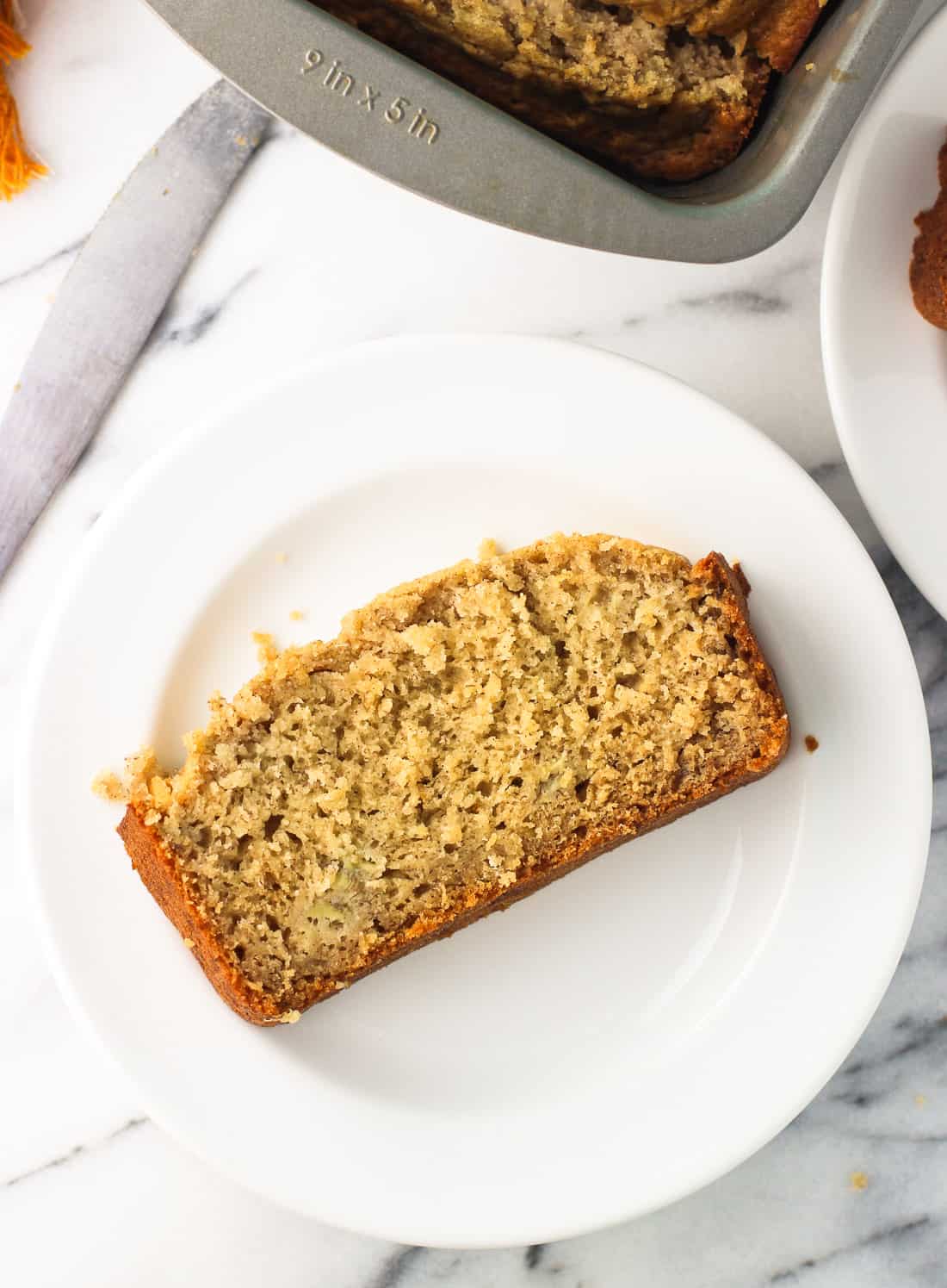 A slice of banana bread on a dessert plate next to the metal loaf pan.