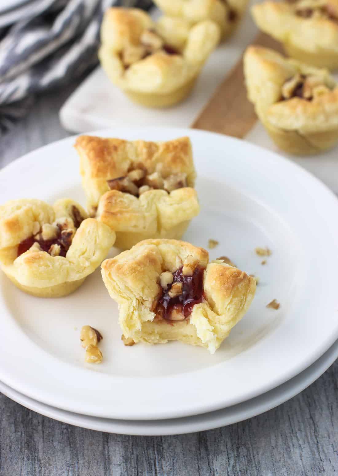 Three baked brie bites, the front one with a bite taken out of it, on an appetizer plate with a tray of them in the background