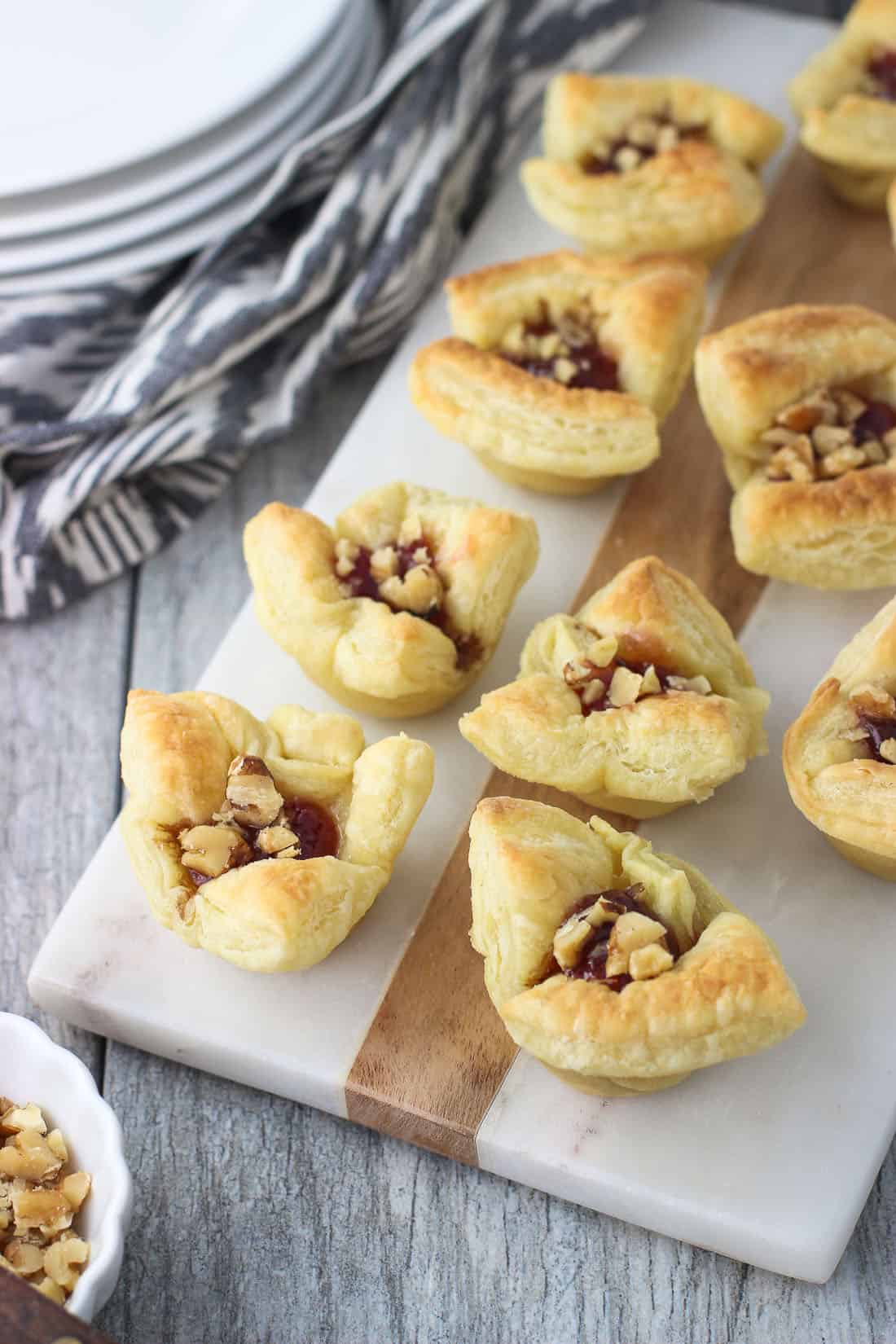 Ten puff pastry brie cups on a marble serving board with a stack of appetizer plates in the background