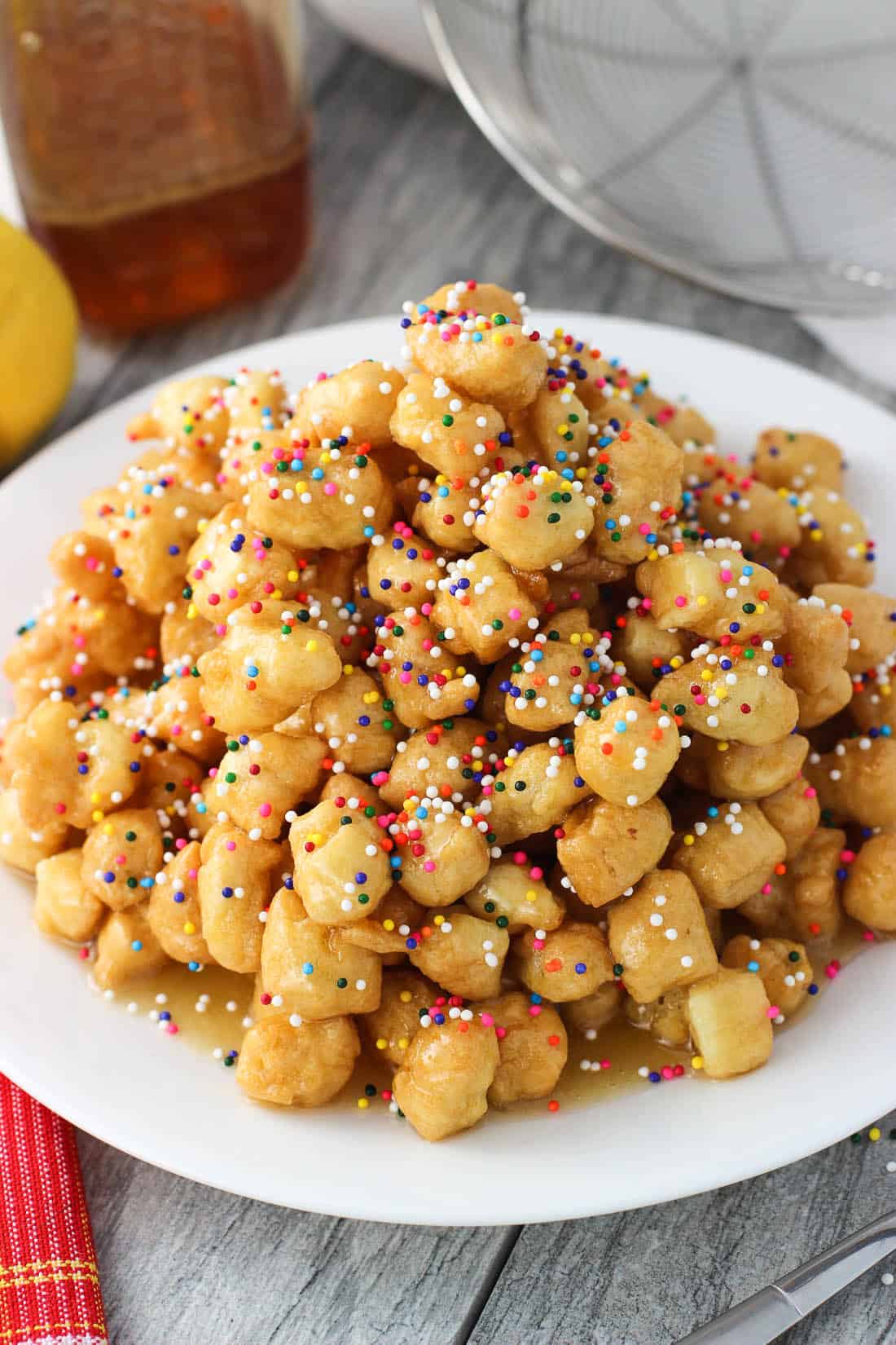 A pile of honey balls on a plate, covered in honey and nonpareil sprinkles