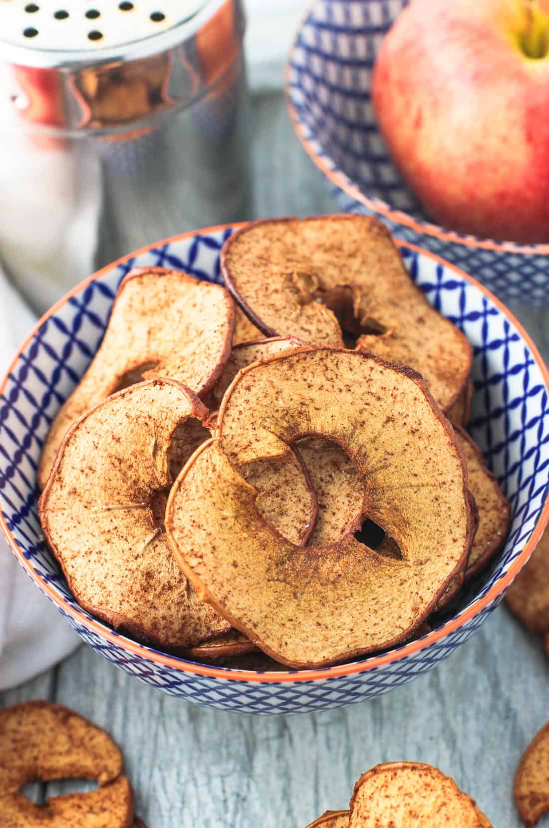 A bowl of apple chip rings next to a metal shaker of cinnamon sugar and a raw apple