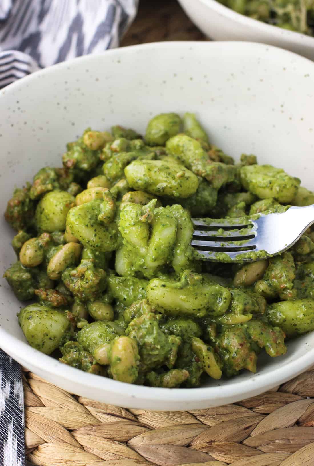 A dish of gnocchi, cannellini beans, and crumbled sausage covered in pesto sauce with a fork spearing several gnocchi