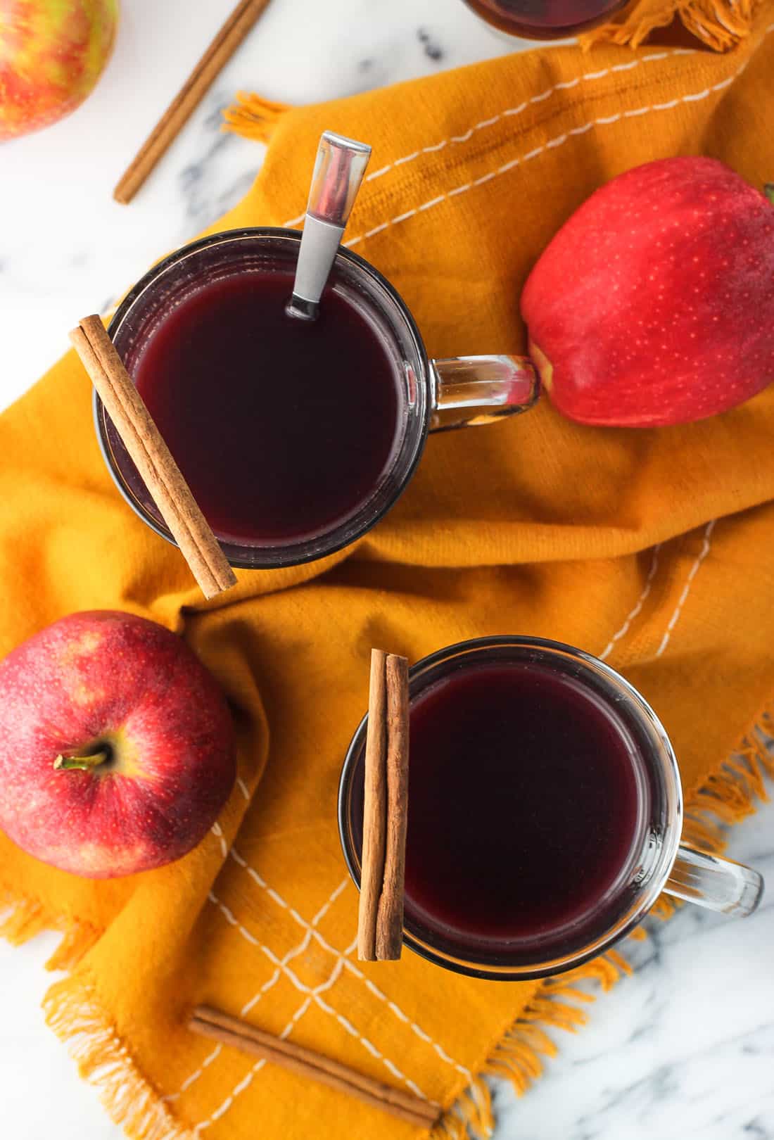 An overhead picture of two mugs of mulled wine garnished with cinnamon sticks. There are fresh apples next to the mugs and one mug has a spoon in it