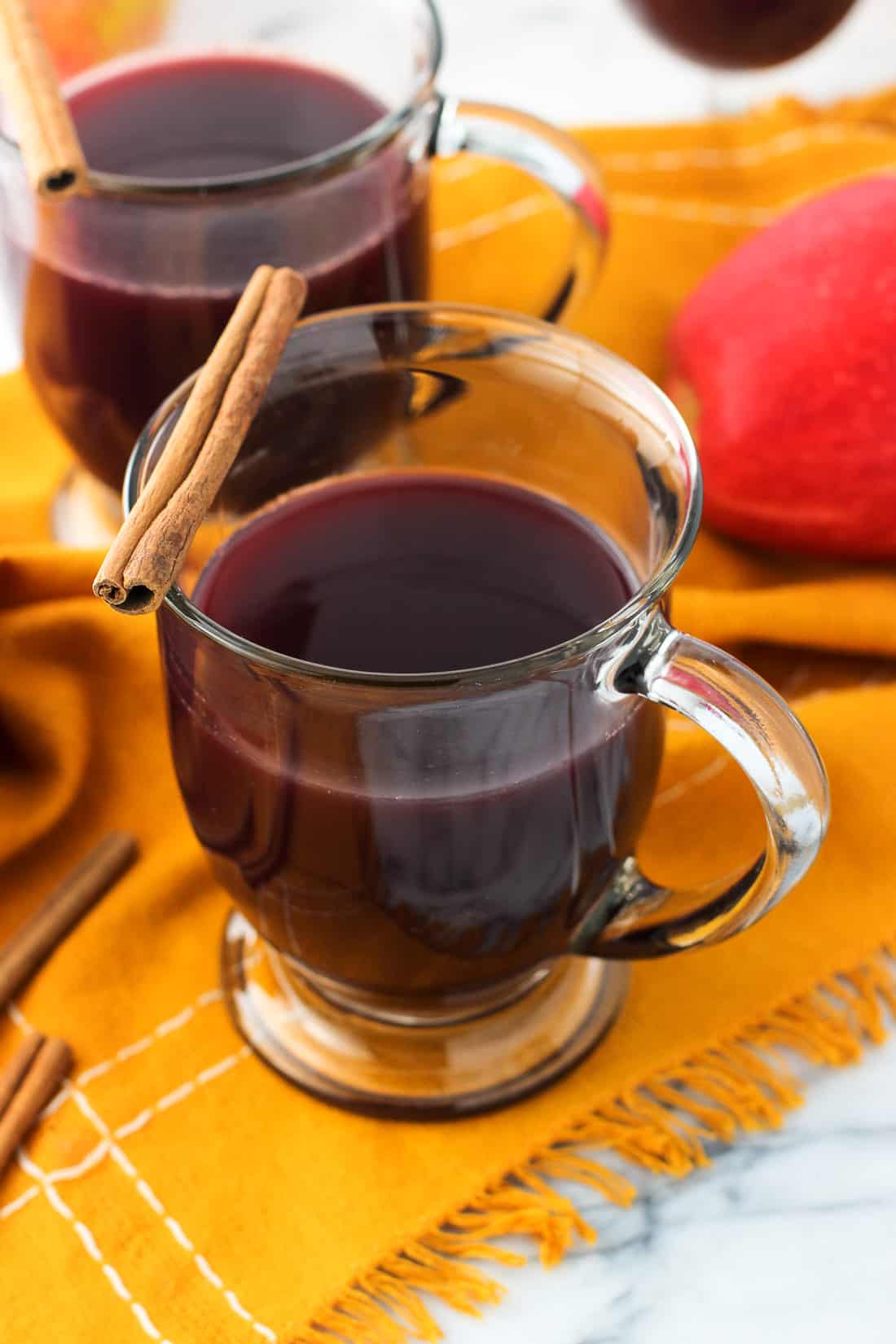 Two clear glass mugs of mulled wine garnished with a cinnamon stick on the rim with an apple in the background.