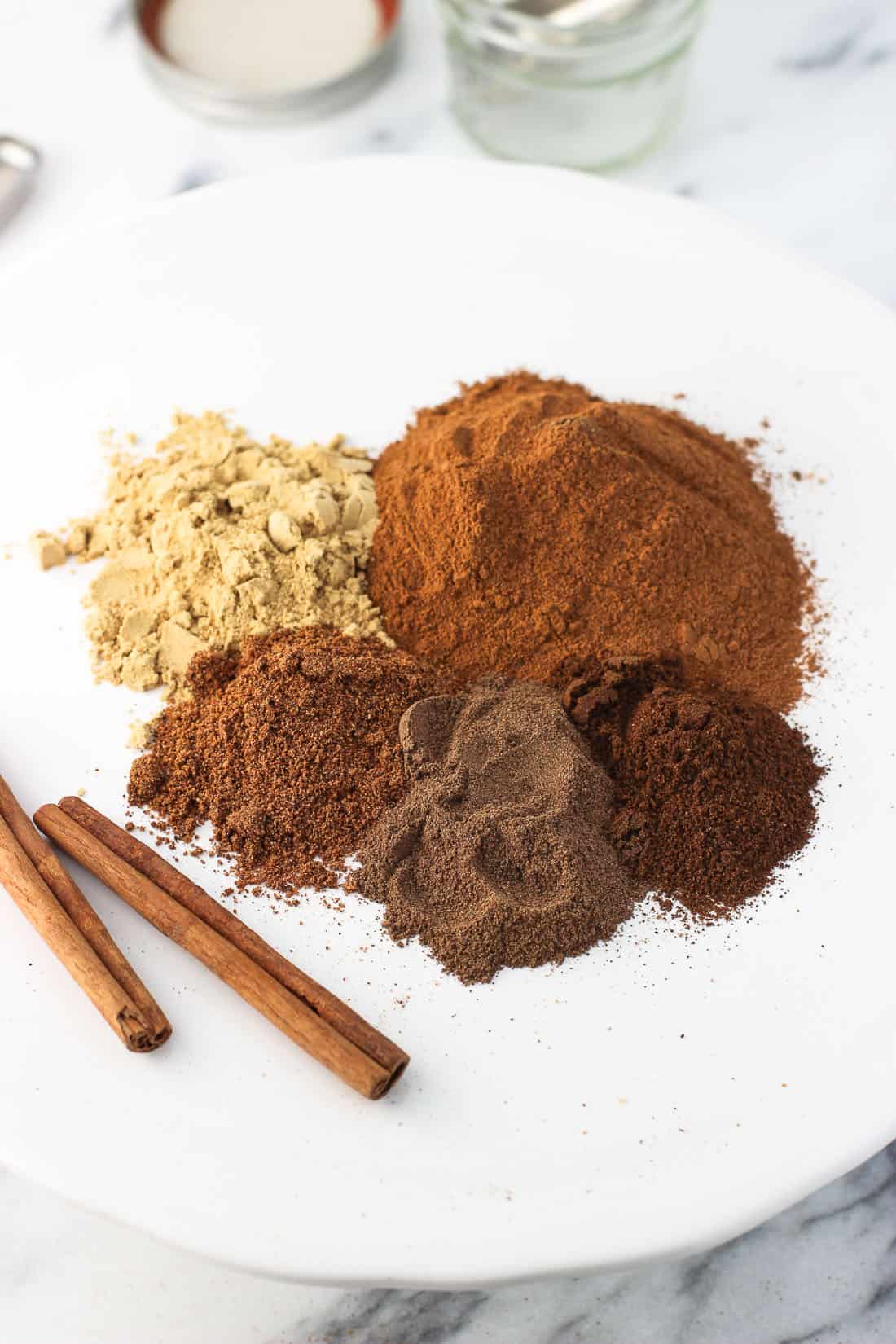 Five piles of ground spices on a ceramic plate next to two whole cinnamon sticks