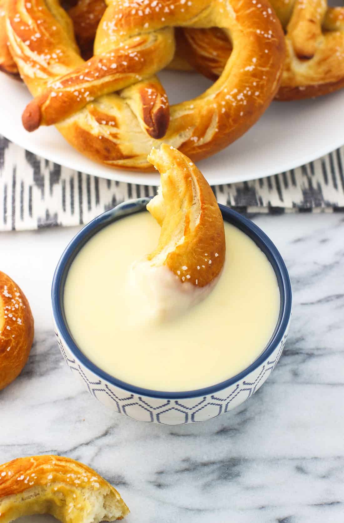 A piece of a soft pretzel dunked in a small bowl of cheese sauce in front of a large plate of pretzels