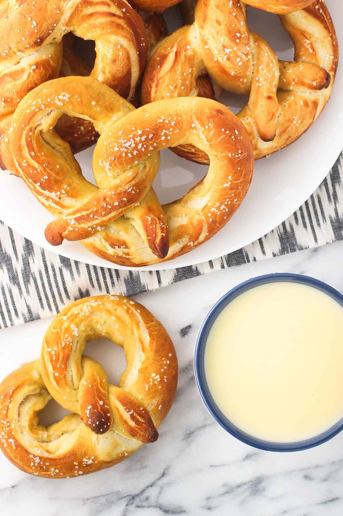 An overhead picture of a plate of pretzels on a marble board next to a small ceramic bowl of cheese sauce for dipping