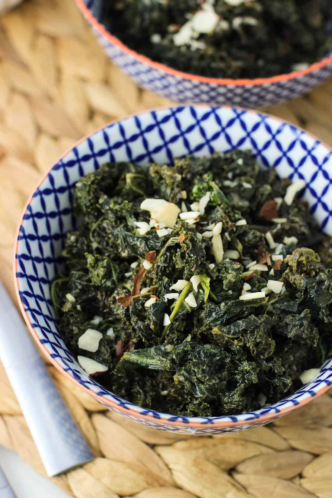 A small ceramic bowl filled with sauteed kale topped with sliced almonds, with another bowl in the background