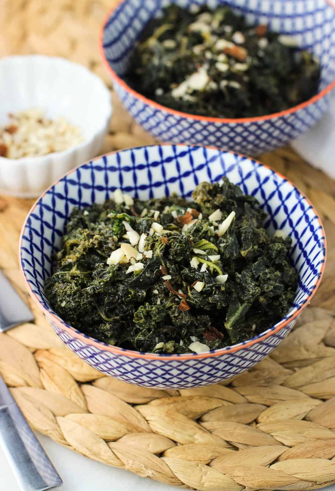 Two bowls of sauteed kale topped with sliced almonds on a woven placemat