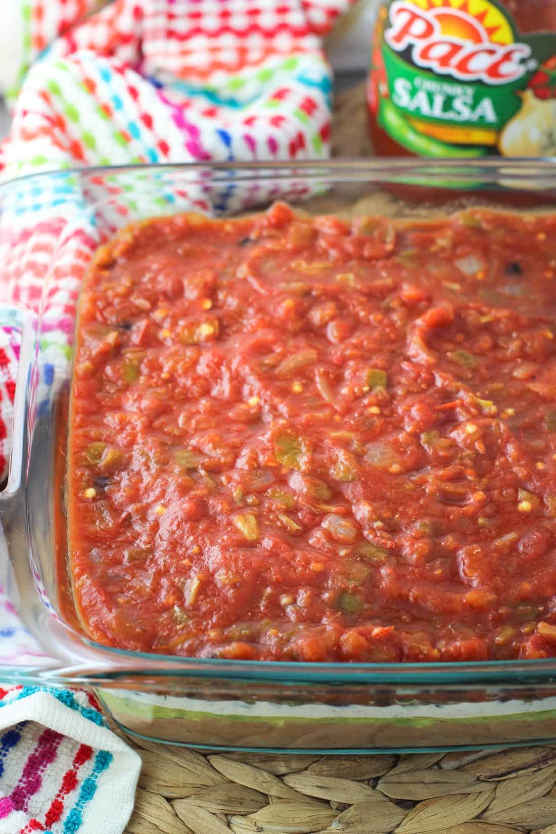 Seven layer dip in a glass baking dish, with the salsa layer on top.