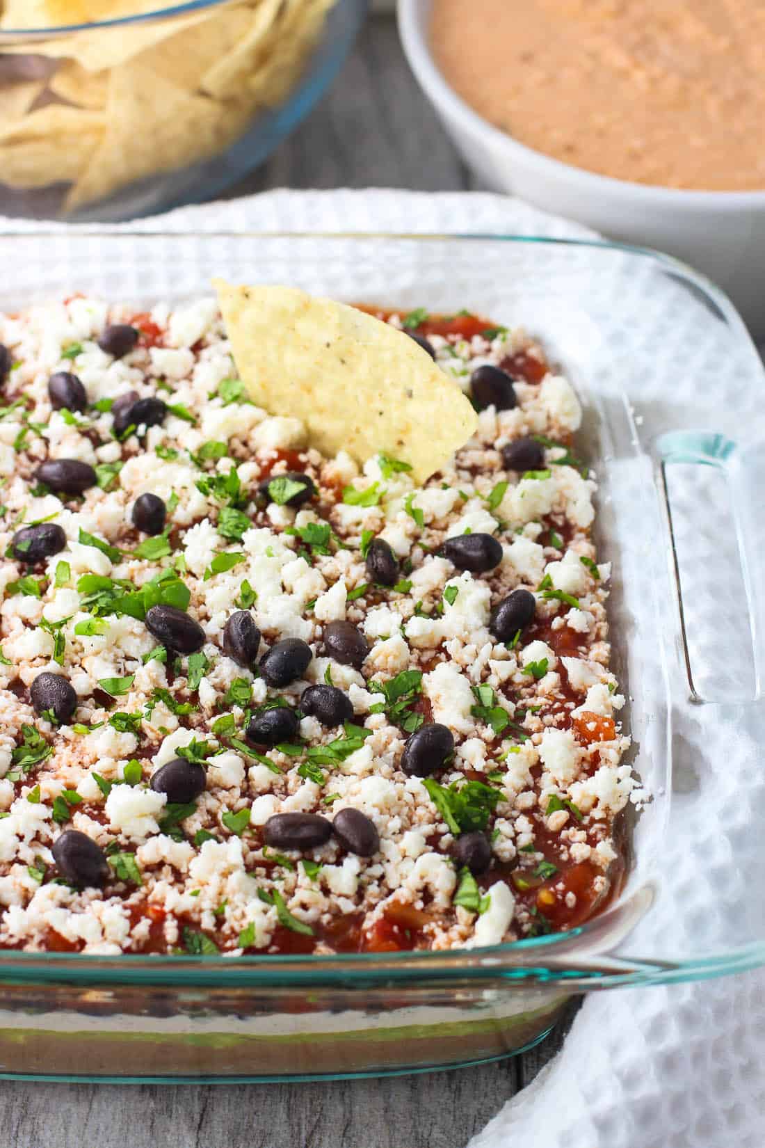 A tortilla chip dunked in the seven layer dip with bowls of tortilla chips and the other blended dip in the background.