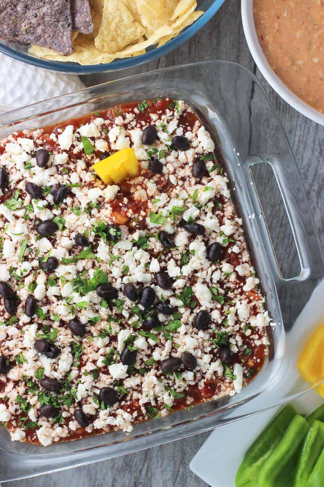 An overhead picture of seven layer dip in a clear glass 8"x8" dish next to a bowl of hot blended dip and a bowl of tortilla chips