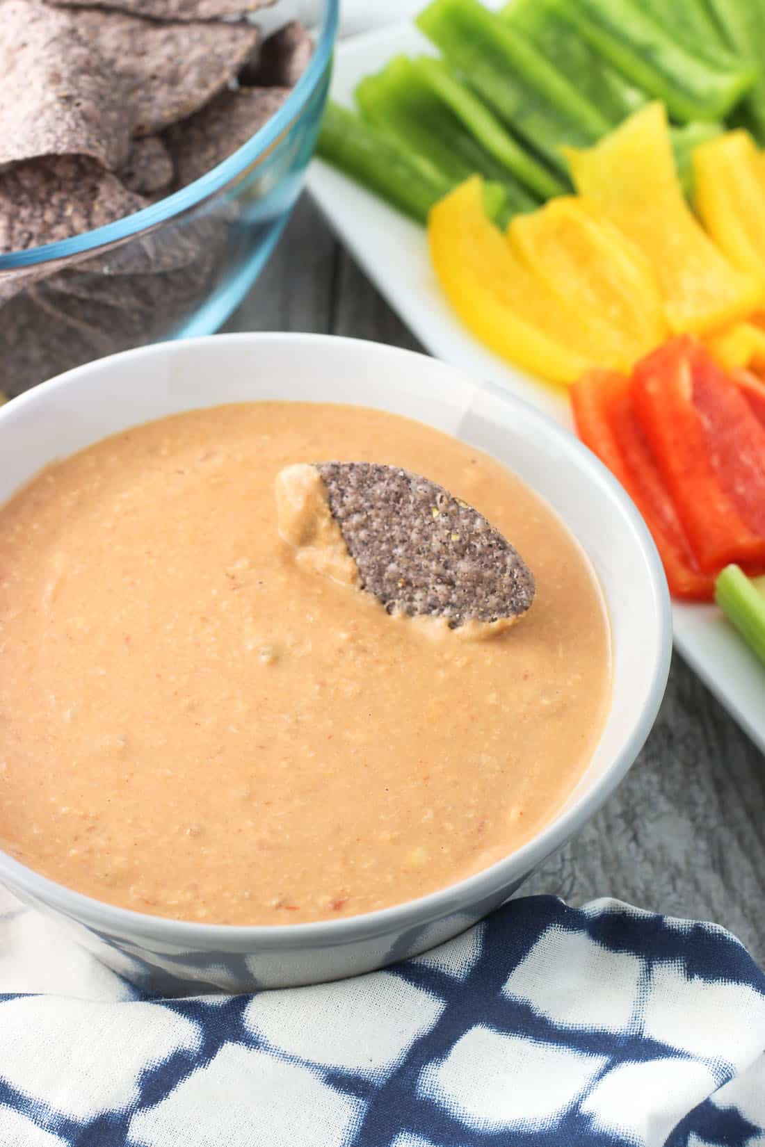 A bowl of warm blended bean dip with a tortilla chip dunked in, in front of a bowl of tortilla chips and a platter of sliced bell peppers