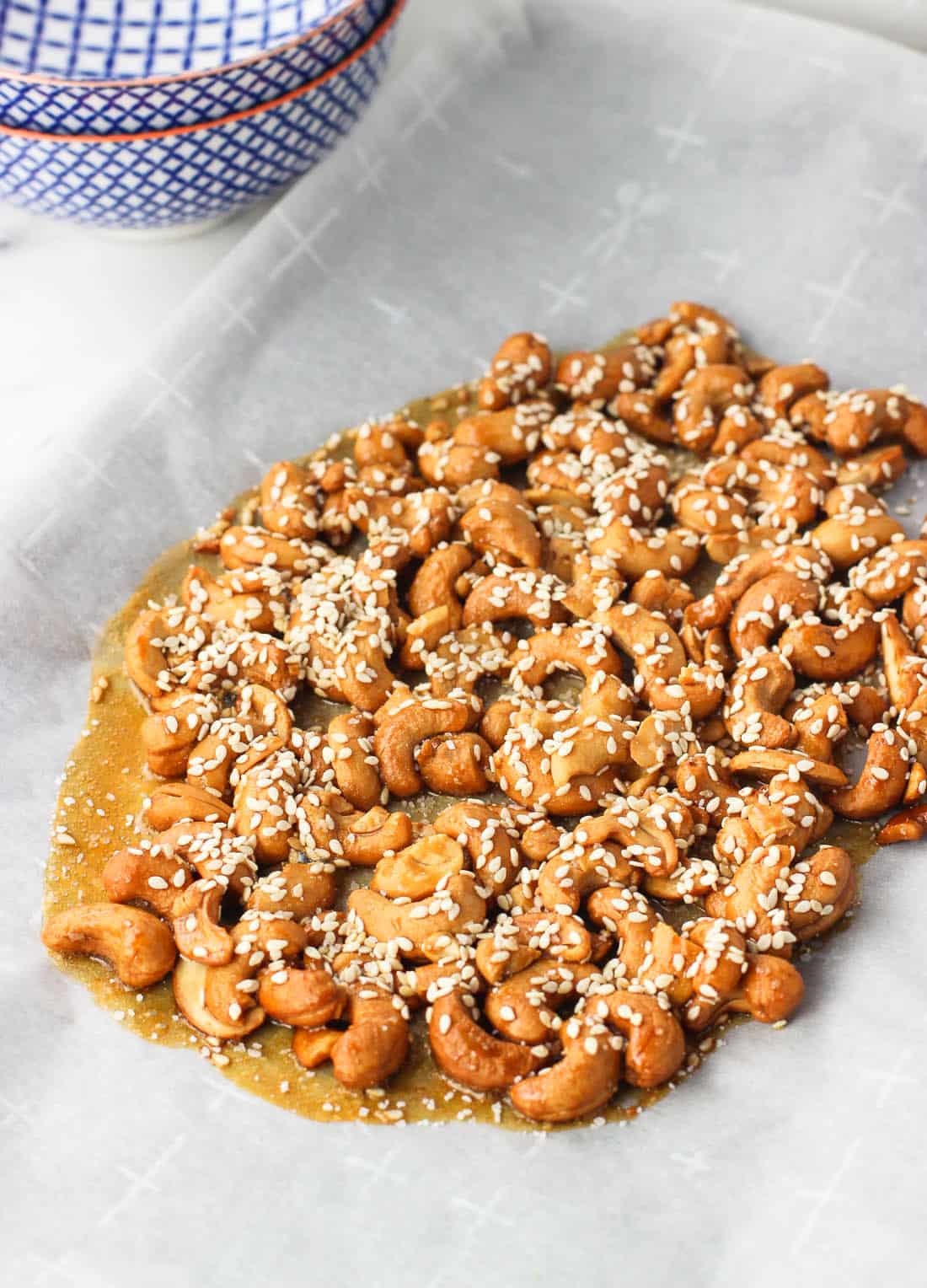 Cashews covered in a honey mixture on a parchment-lined sheet pan, topped with sesame seeds before breaking apart into clusters