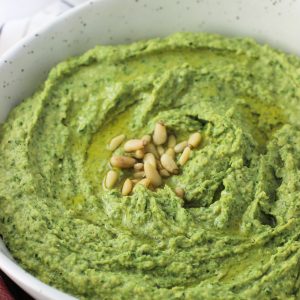 A serving bow of kale pesto hummus topped with olive oil and pine nuts.