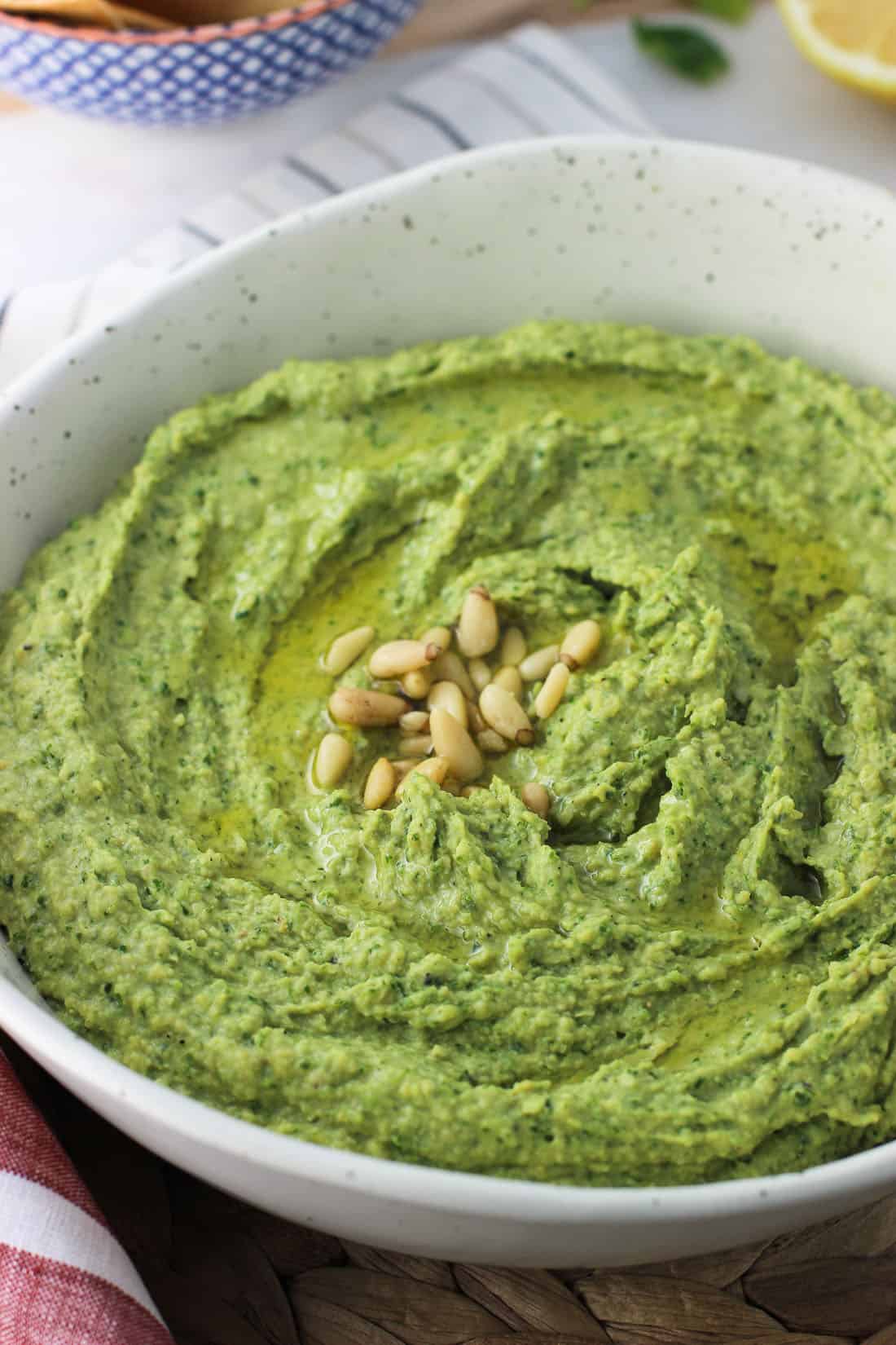 A serving bow of kale pesto hummus topped with olive oil and pine nuts.