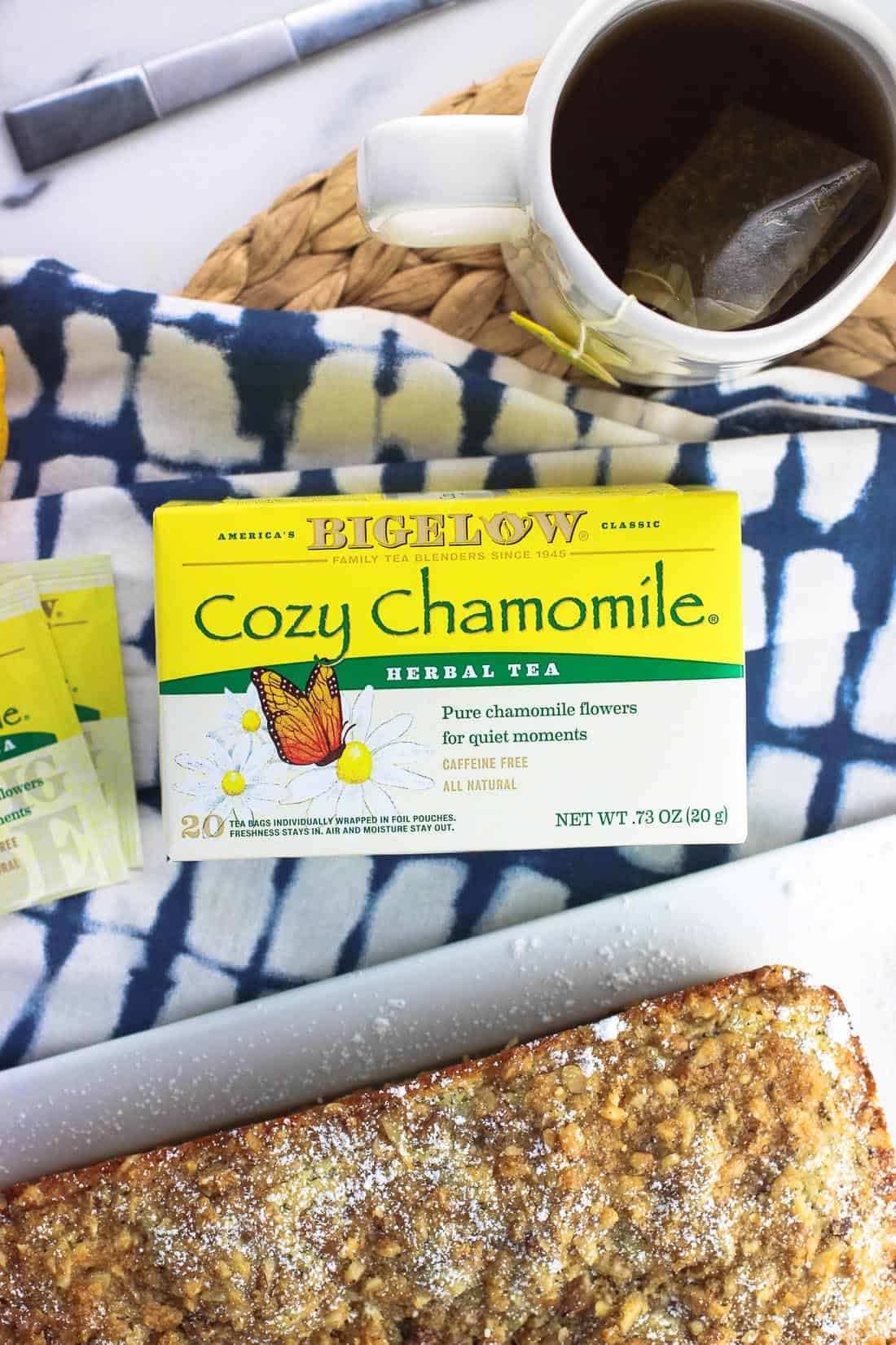 An overhead picture of a box of Bigelow Cozy Chamomile tea next to a mug of tea, two foil-wrapped teabags, and the bread