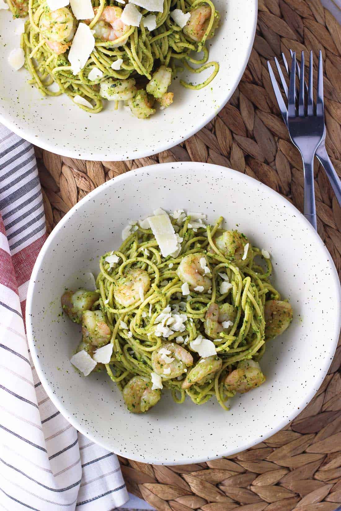 An overhead shot of two shallow dinner dishes of pasta tossed in pesto sauce with shrimp and shaved Parmesan.