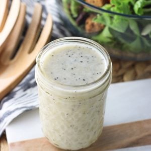 A glass jar filled with dressing in front of a bowl fo salad and wooden tongs