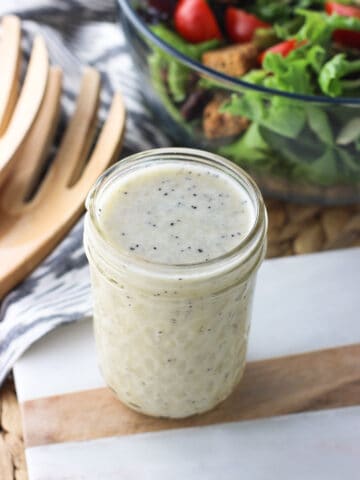 A glass jar filled with dressing in front of a bowl fo salad and wooden tongs